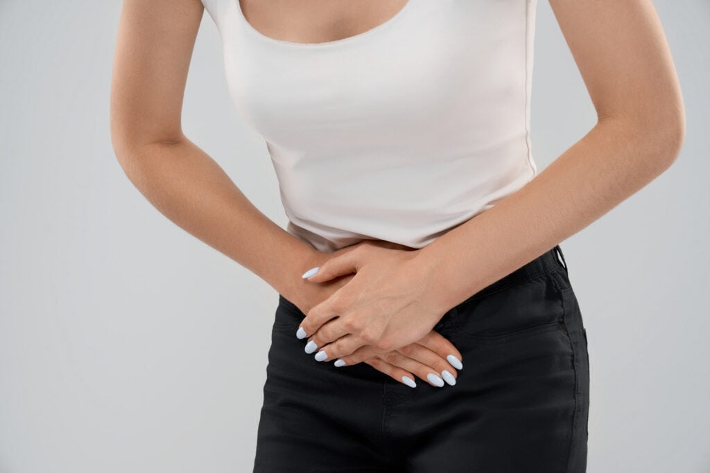 Clolse up of bending incognito caucasian woman in casual suit posing over gray isolated background and touching lower abdomen because of ache. Front crop of brunette suffering from pain in stomach.